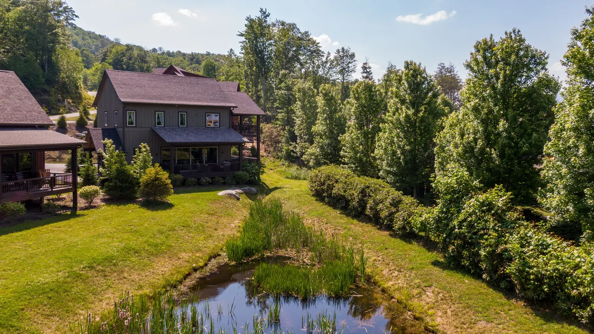 This image showcases a tranquil exterior view of the house nestled beside a picturesque pond, surrounded by mature trees and lush greenery. The calm water reflects the sky, and the porch offers a welcoming spot for morning coffee or quiet moments together in nature.