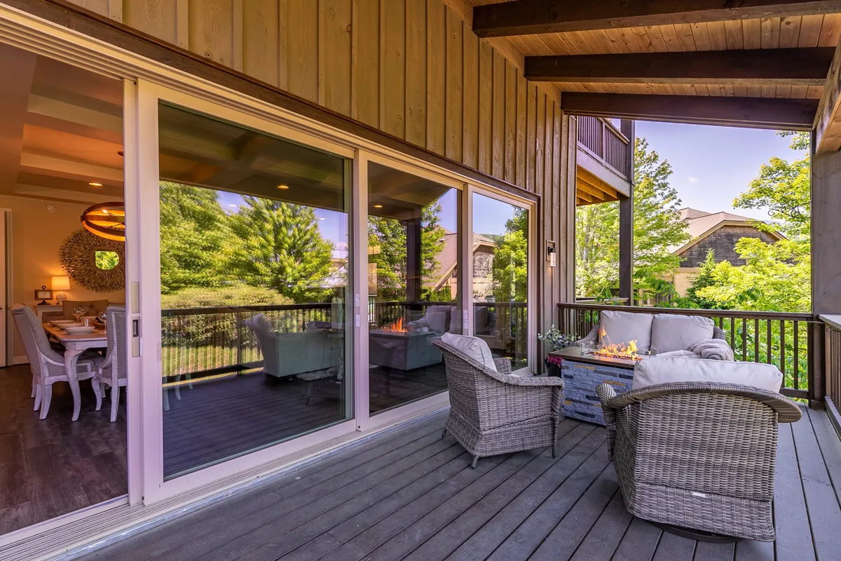 This image shows a comfortable outdoor seating area with wicker chairs arranged around a fire pit, set against green landscaping and large windows of the home. The inviting space is perfect for morning coffee or evening relaxation with family and friends.