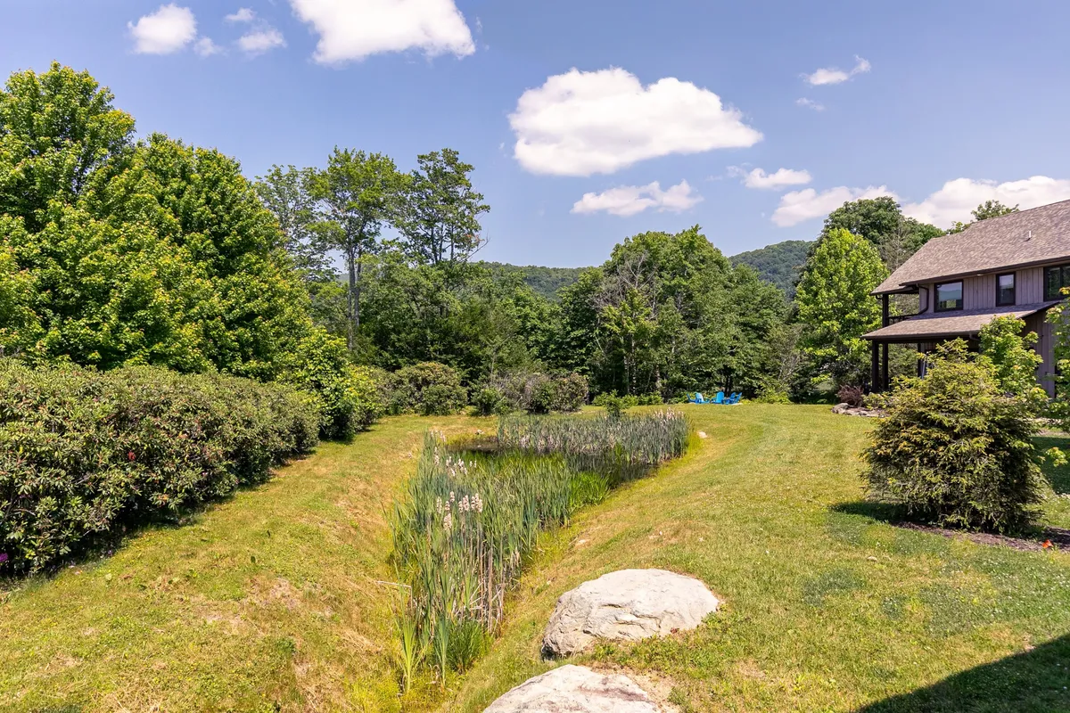 A serene exterior shot featuring a house nestled among lush greenery, a tranquil pond with large rocks, and open space ideal for family nature walks or relaxing by the water. The scene conveys a sense of adventure and peacefulness in a natural setting.