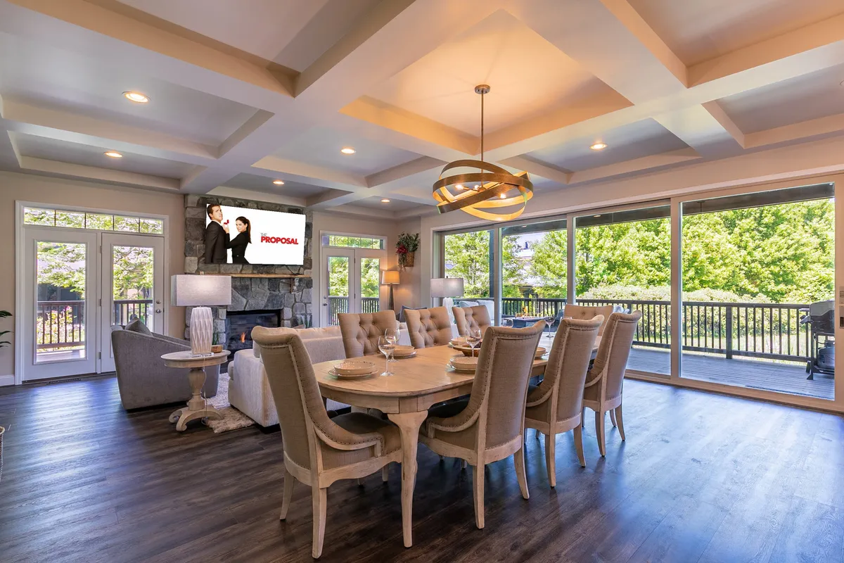 A spacious, inviting living area with a dining table, elegant chandeliers, television, large windows, and a cozy fireplace. The view through the windows hints at the outdoors, and the fireplace provides a perfect spot to relax after a day outside.