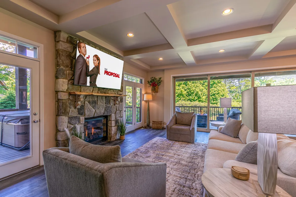 A cozy living room with a prominent stone fireplace, a large window framing natural light, and a TV screen displaying a movie. Plush seating forms an inviting gathering spot, and the ambiance is both warm and welcoming.