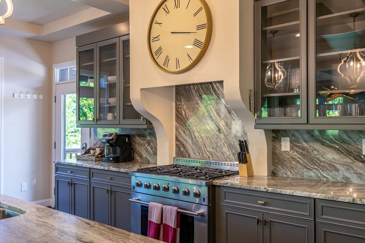 A stylish, modern kitchen featuring a large wall clock, a prominent gas stove with marble backsplash, a generous kitchen island, and ample space for preparing meals. The area is bright and welcoming, perfect for culinary activities.
