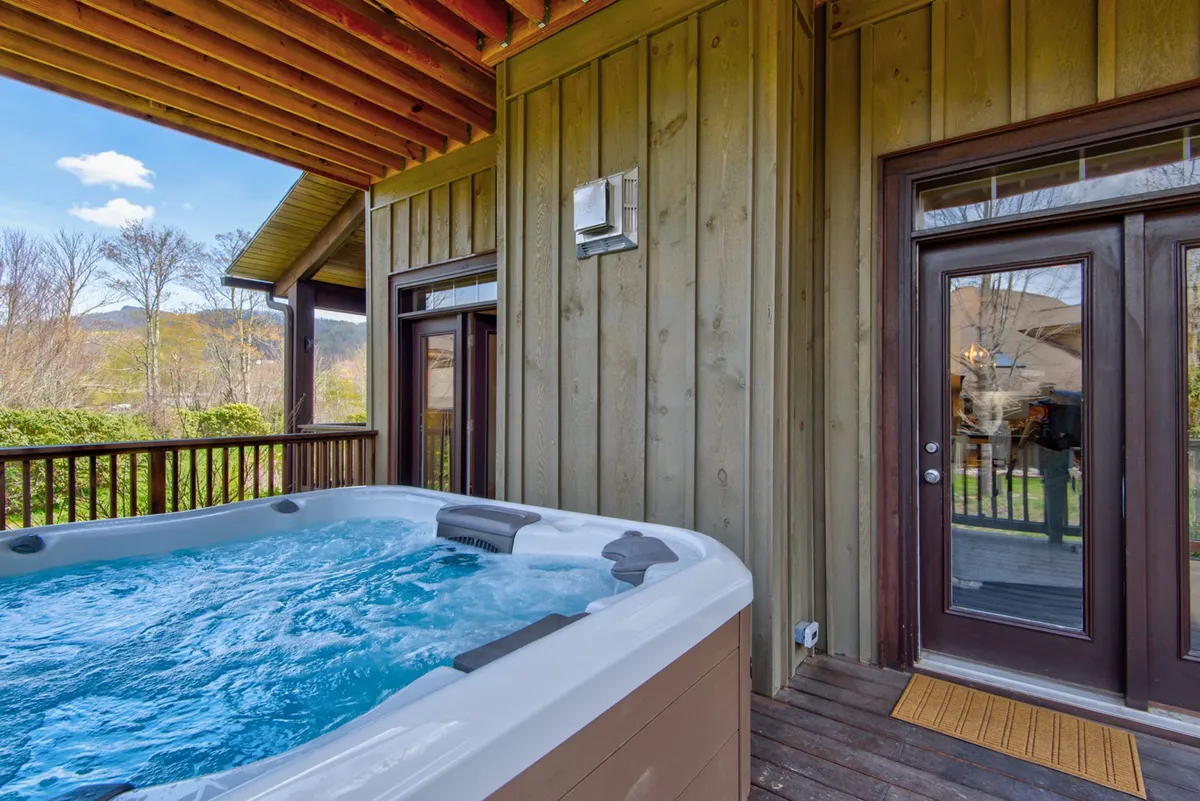 This exterior view features a private wooden deck with a bubbling hot tub and door access to the house, all set against a backdrop of mountain views. The inviting hot tub is positioned for sunset and stargazing experiences, ideal for romantic evenings.