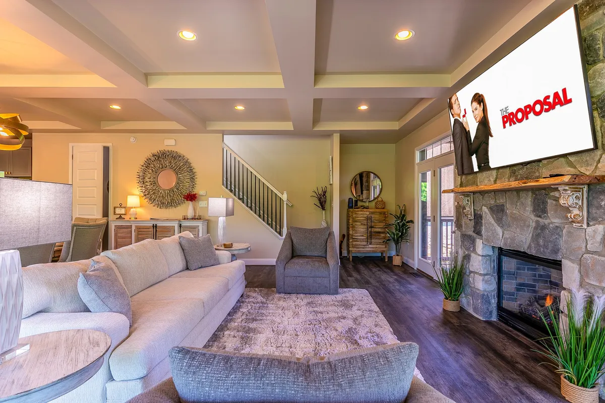 A spacious, open-plan living area with a large sectional sofa, wall-mounted television playing a movie, and a modern fireplace. The layout emphasizes comfort and encourages socializing or relaxing with family.