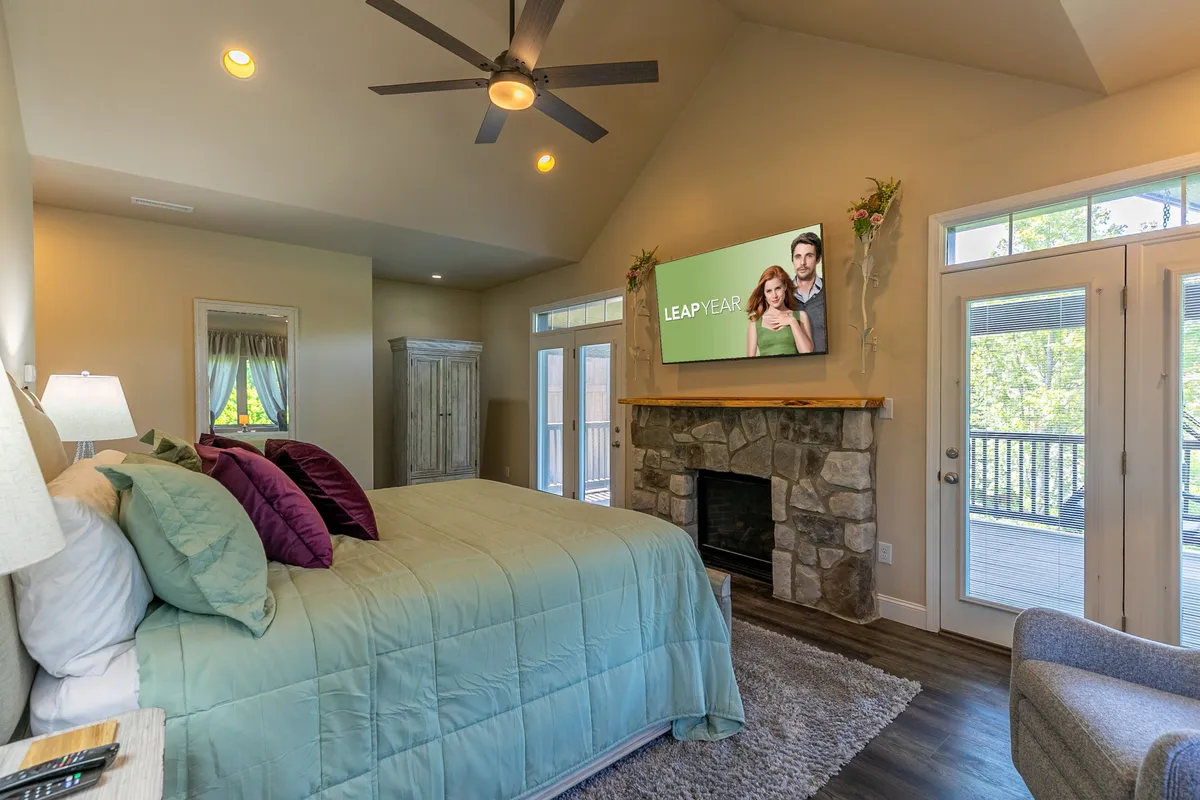 A spacious bedroom featuring a large bed with teal and purple bedding, a stone fireplace, flat-screen TV, and expansive windows flooding the room with natural light. The ambiance is tranquil, blending comfort and luxury, and the presence of a fireplace hints at a cozy, pampering retreat.