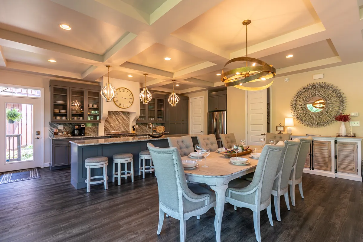 An inviting open-plan area with a dining table set near a kitchen island. Pendant lights hang above the island, and reflective details like a mirror and clock add style. The space is set up for family gatherings or dinner parties, with a natural connection between cooking and dining.
